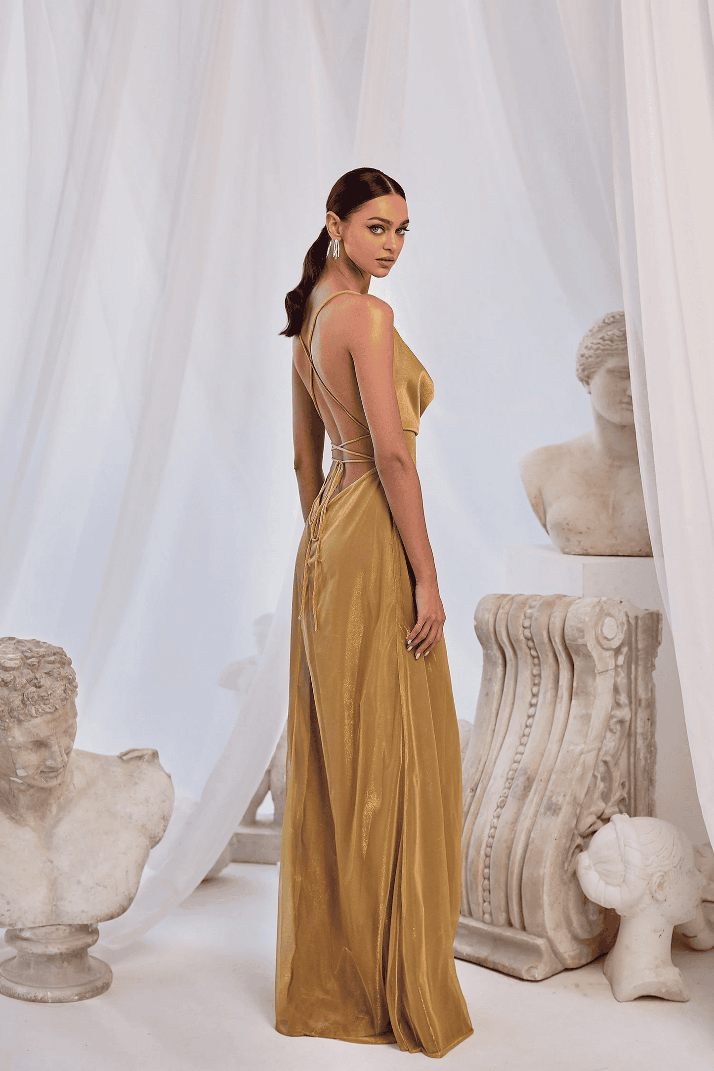 A model in an Annabella gold satin gown standing in front of a white curtain
